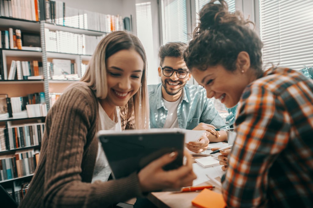 Three students smiling and using a tablet together in a library